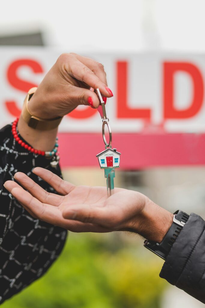 pexels photo 8293717 8293717 Close-up of hands exchanging house key with colorful SOLD sign in the background.